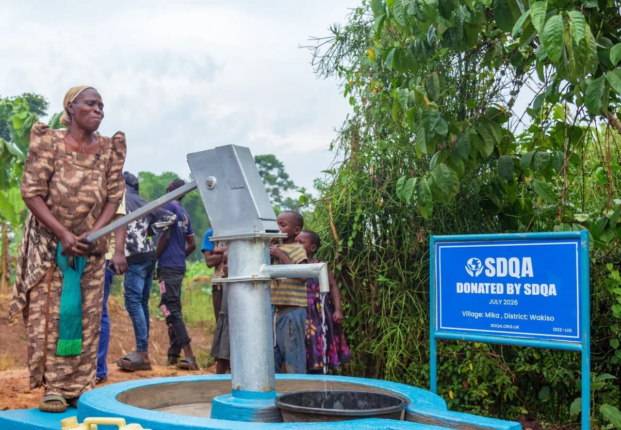 A woman stands beside the finished hand pump at Mika, with children at the spout and the blue SDQA sign behind.